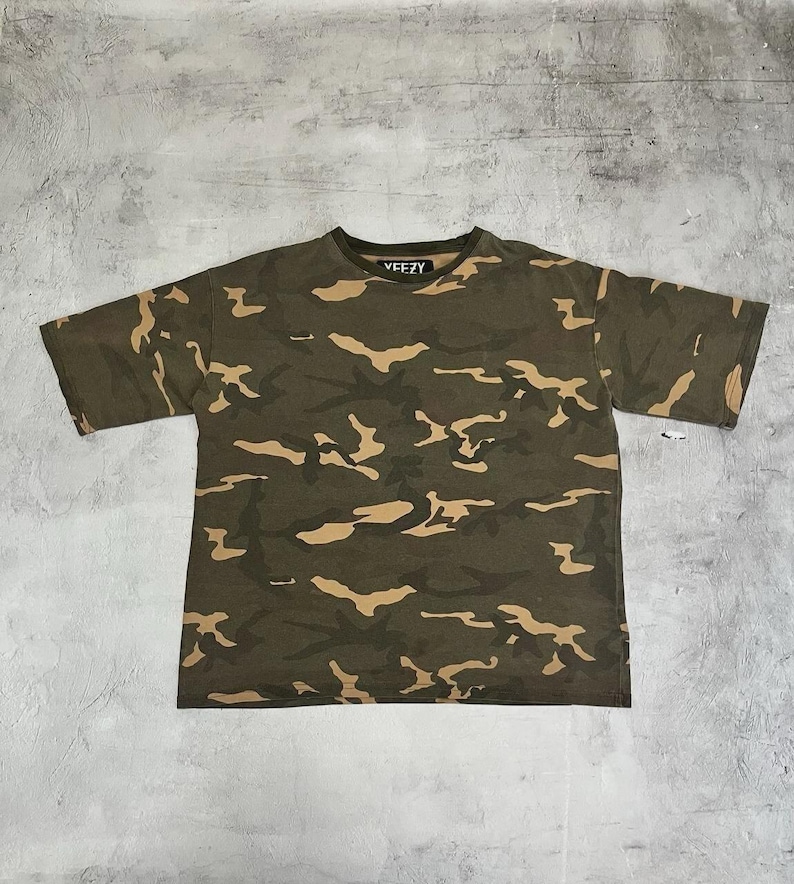 Puede incluir: Camiseta de camuflaje verde oliva con cuello redondo y mangas cortas. La camiseta presenta un estampado de formas marrones y beige. La palabra "YEEZY" est&aacute; impresa en blanco en el cuello.
