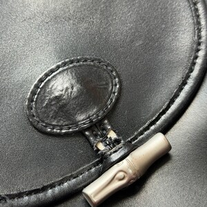May include: Close-up of a black leather handbag. The bag features a silver-toned bamboo-style clasp and a stitched oval detail. The leather has a slight sheen, and the stitching is visible along the edges. The bag is a classic style.