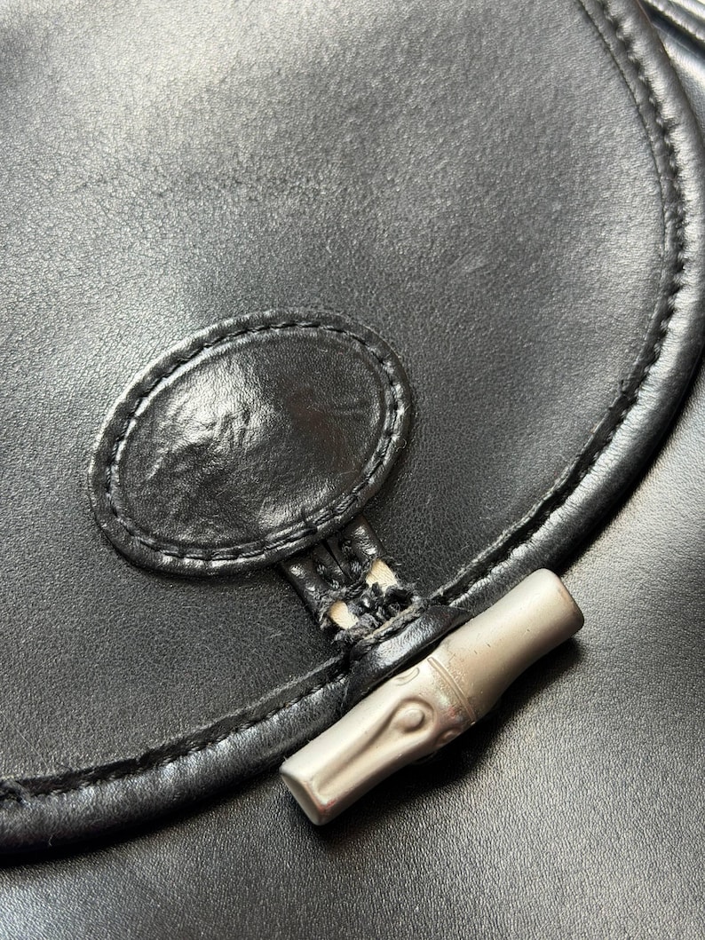 May include: Close-up of a black leather handbag. The bag features a silver-toned bamboo-style clasp and a stitched oval detail. The leather has a slight sheen, and the stitching is visible along the edges. The bag is a classic style.