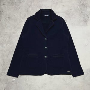 Puede incluir: Un blazer azul marino de diseño clásico. La chaqueta presenta solapas con muescas, dos bolsillos delanteros y tres botones plateados. El blazer tiene un corte entallado y está hecho de una tela suave y texturizada.