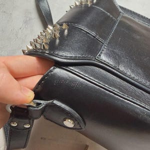 Puede incluir: Bolso de hombro de cuero negro con tachuelas c&oacute;nicas plateadas en el borde superior. El bolso tiene un cierre de solapa y un cierre de metal plateado. La correa es negra y el bolso parece estar hecho de cuero liso.