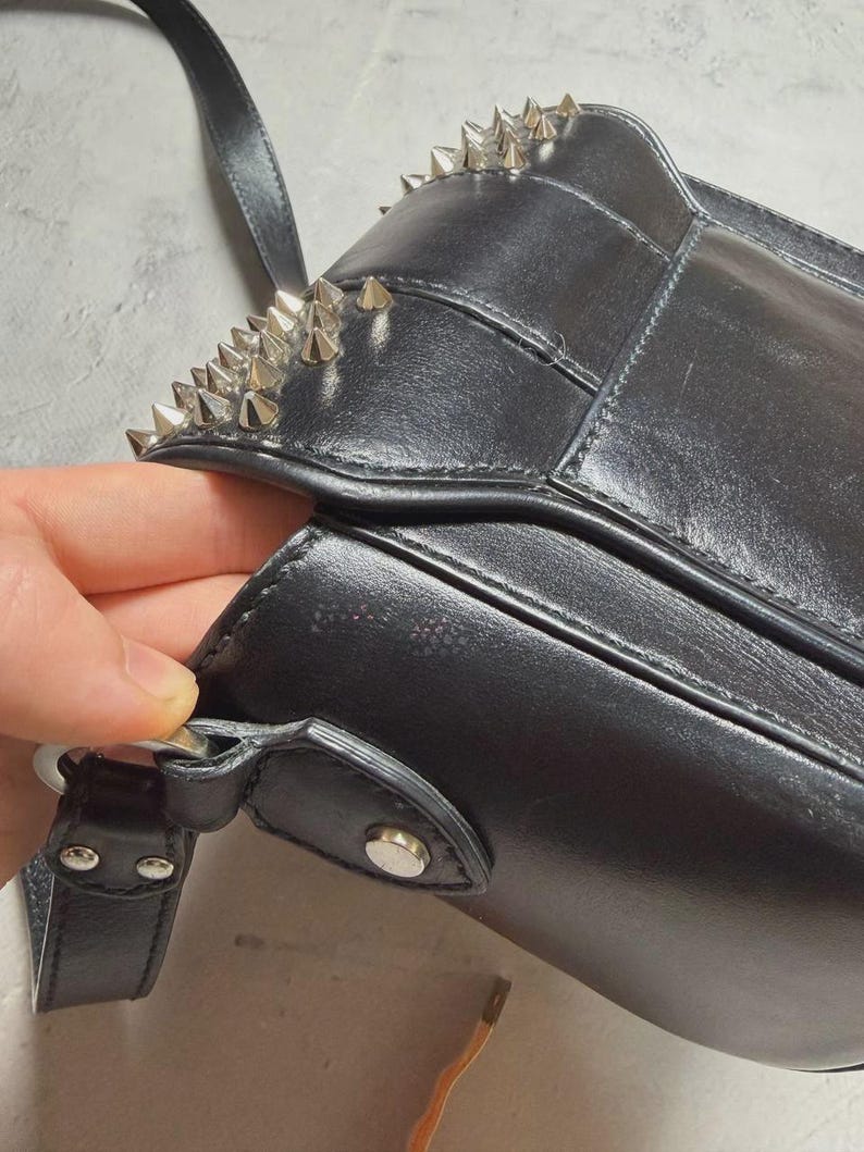 Puede incluir: Bolso de hombro de cuero negro con tachuelas c&oacute;nicas plateadas en el borde superior. El bolso tiene un cierre de solapa y un cierre de metal plateado. La correa es negra y el bolso parece estar hecho de cuero liso.