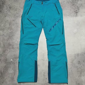 May include: Teal-colored Dynafit ski pants with multiple zippered pockets and the brand name printed on the leg. The pants have a button and zipper closure at the waist and reinforced panels at the knees and lower legs. The pants are designed for outdoor winter activities.