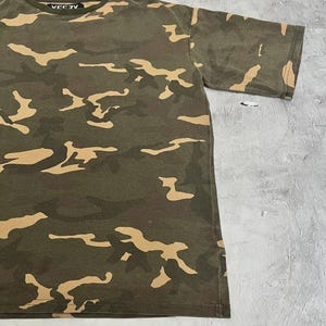 Puede incluir: Una camiseta de camuflaje verde oliva con formas irregulares de color beige. La camiseta tiene mangas cortas y cuello redondo con la palabra "VEEZY" impresa en blanco en el cuello.