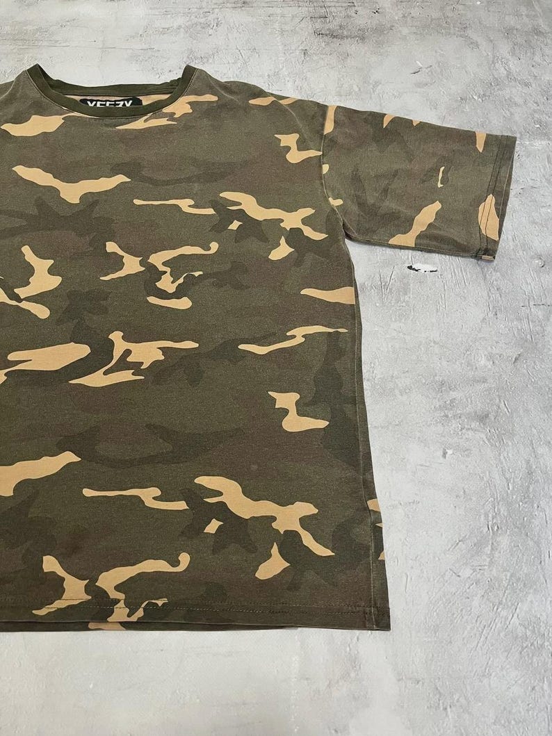 Puede incluir: Una camiseta de camuflaje verde oliva con formas irregulares de color beige. La camiseta tiene mangas cortas y cuello redondo con la palabra "VEEZY" impresa en blanco en el cuello.