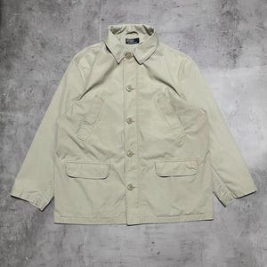 May include: A light tan jacket with a button-up front, two front pockets, and a collared neckline. The jacket has a relaxed fit and is made of a lightweight material. The Polo logo is visible on the collar.