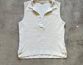 May include: A sleeveless, off-white collared shirt. The shirt has a V-neck and a textured fabric. The brand name is visible on the collar. The shirt is laid flat on a gray surface.