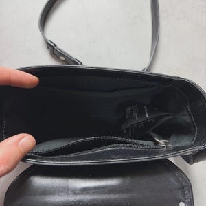 Puede incluir: Bolso bandolera de cuero negro con una correa larga. El bolso tiene un cierre de solapa con tachuelas plateadas. El interior es visible, mostrando un bolsillo con cremallera y una etiqueta. El nombre de la marca es visible.