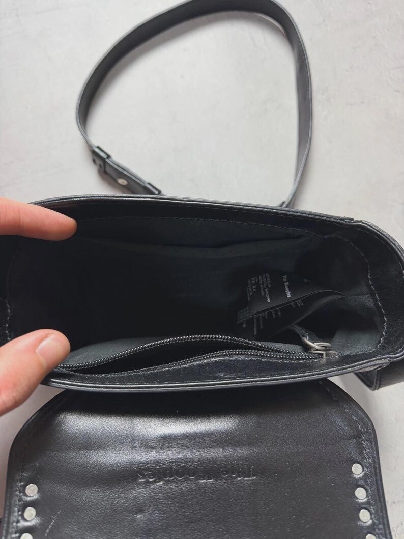 Puede incluir: Bolso bandolera de cuero negro con una correa larga. El bolso tiene un cierre de solapa con tachuelas plateadas. El interior es visible, mostrando un bolsillo con cremallera y una etiqueta. El nombre de la marca es visible.