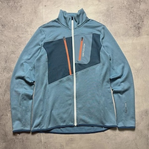 May include: A light blue zip-up jacket with a high collar. The jacket features a teal chest pocket with an orange zipper and the brand name "ORTOVOX" printed vertically. The jacket has a textured pattern and a white zipper.