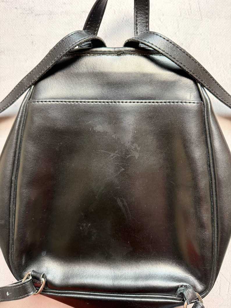May include: A black leather backpack with adjustable straps. The backpack has a small pocket on the back and silver-colored hardware. The leather shows some wear and tear, with visible scratches and marks on the surface.