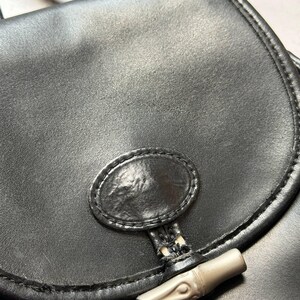 May include: Close-up of a black leather handbag with a curved flap. The bag features a silver-toned bamboo-style clasp and a stitched oval detail on the front. The leather has a slightly textured appearance.