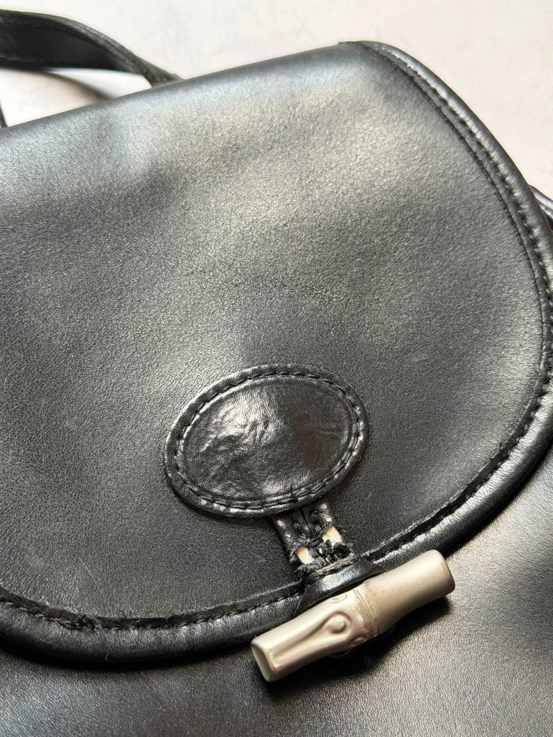 May include: Close-up of a black leather handbag with a curved flap. The bag features a silver-toned bamboo-style clasp and a stitched oval detail on the front. The leather has a slightly textured appearance.