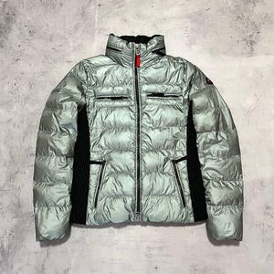 May include: A light green puffer jacket with a high collar and a full-length zip. The jacket has black panels on the sides and around the pockets. The zip pull is red. This winter coat is designed for warmth and style.