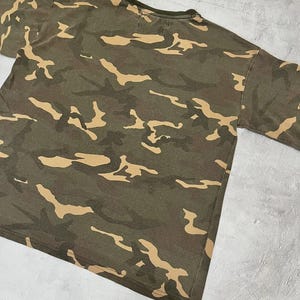 Puede incluir: Una camiseta con estampado de camuflaje verde oliva con formas irregulares de color beige. La camiseta tiene cuello redondo y mangas cortas. La tela parece ser un material suave y desgastado.