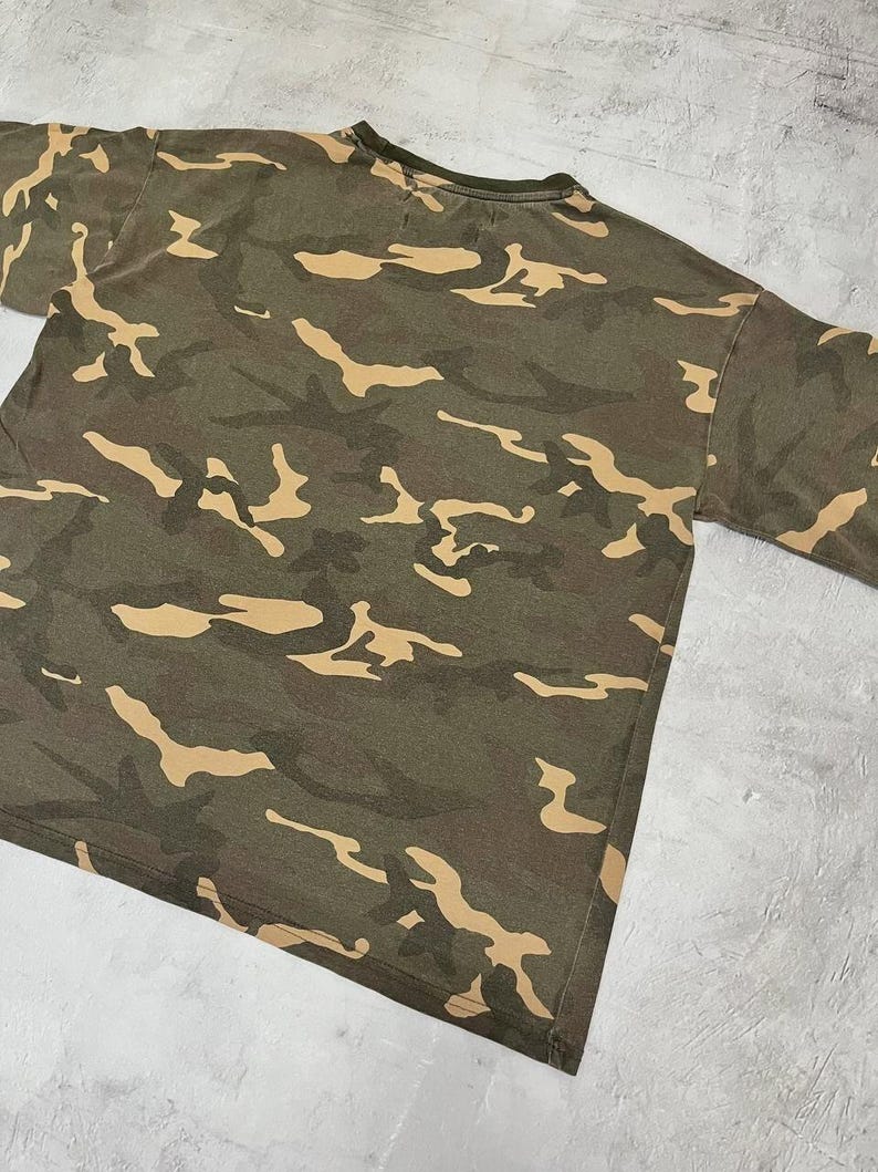 Puede incluir: Una camiseta con estampado de camuflaje verde oliva con formas irregulares de color beige. La camiseta tiene cuello redondo y mangas cortas. La tela parece ser un material suave y desgastado.