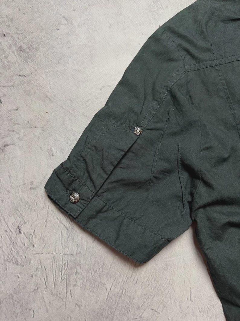 May include: Dark green short-sleeved shirt with a buttoned cuff. The shirt has a classic design with a button on the sleeve and a decorative button near the shoulder. The fabric appears to be a lightweight material.
