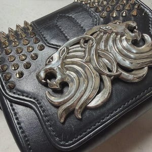 Puede incluir: Bolso bandolera de cuero negro con emblema de cabeza de le&oacute;n plateado y detalles de tachuelas. El bolso tiene cierre de solapa y una correa larga. Tama&ntilde;o peque&ntilde;o.