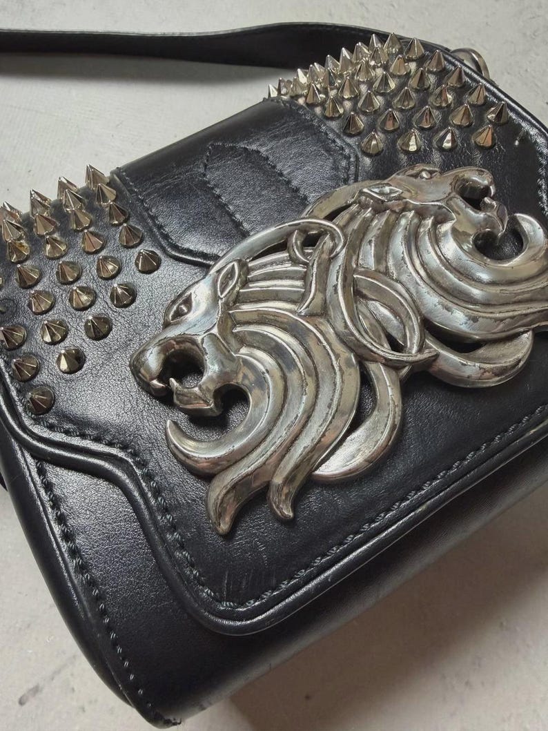 Puede incluir: Bolso bandolera de cuero negro con emblema de cabeza de le&oacute;n plateado y detalles de tachuelas. El bolso tiene cierre de solapa y una correa larga. Tama&ntilde;o peque&ntilde;o.