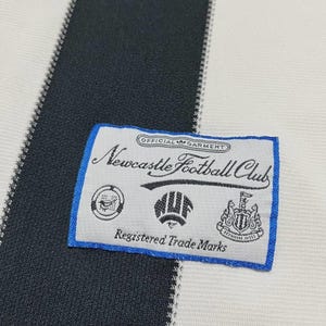 Puede incluir: Primer plano de una tela a rayas blancas y negras con una etiqueta con borde azul. La etiqueta dice "Newcastle Football Club" con varios emblemas y el texto "Registered Trade Marks".