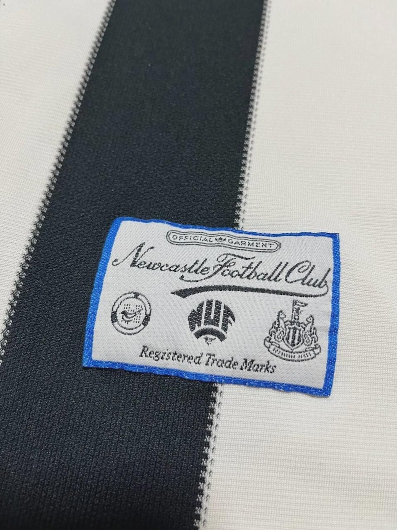 Puede incluir: Primer plano de una tela a rayas blancas y negras con una etiqueta con borde azul. La etiqueta dice "Newcastle Football Club" con varios emblemas y el texto "Registered Trade Marks".