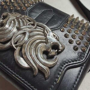 Puede incluir: Bolso de hombro de cuero negro con emblema de cabeza de le&oacute;n plateado y tachuelas. El bolso tiene un cierre de solapa y una correa larga. La cabeza de le&oacute;n plateada es un elemento decorativo prominente.