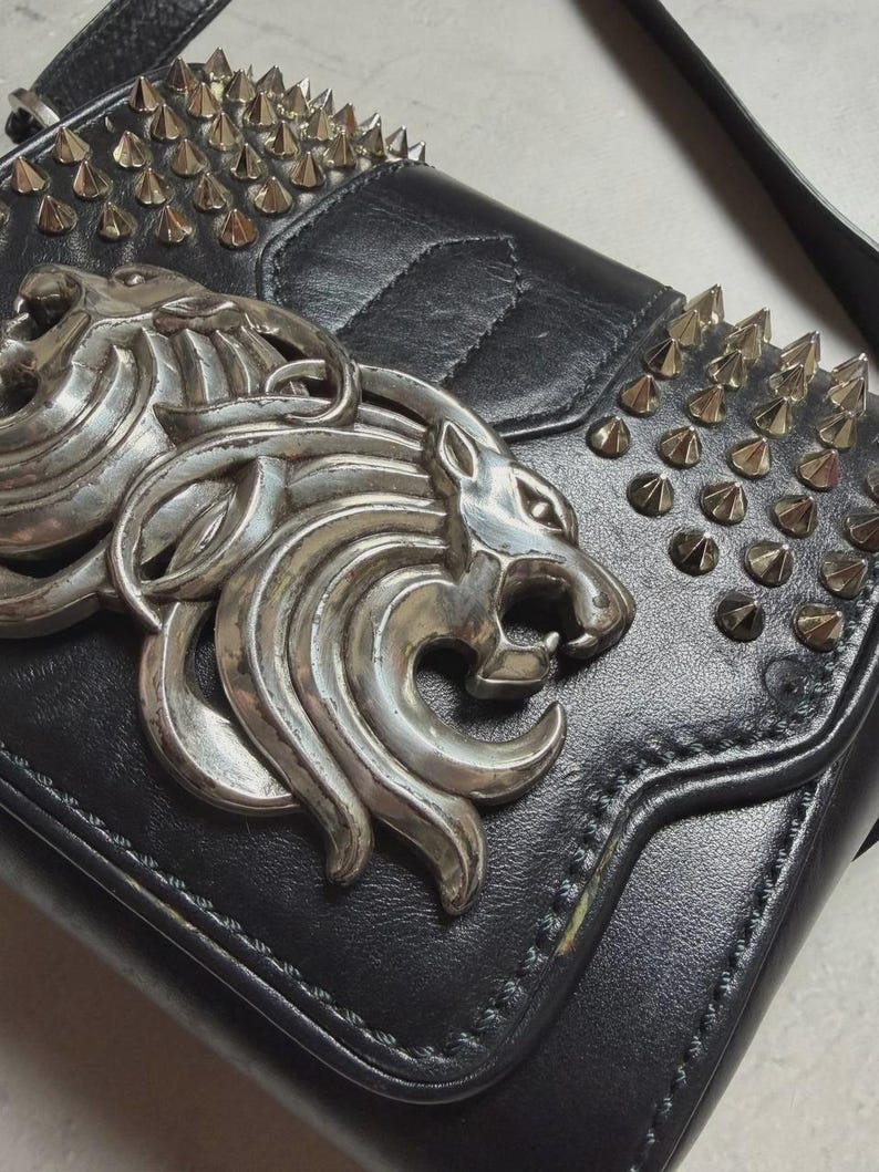 Puede incluir: Bolso de hombro de cuero negro con emblema de cabeza de le&oacute;n plateado y tachuelas. El bolso tiene un cierre de solapa y una correa larga. La cabeza de le&oacute;n plateada es un elemento decorativo prominente.