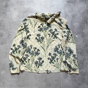May include: A cream-coloured long-sleeved blouse featuring a floral design of blue flowers and green stems. The blouse has a bow detail at the neckline.