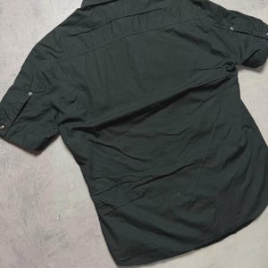 May include: Dark green short-sleeved shirt with a button-down collar. The shirt has two chest pockets and buttoned sleeve tabs. The back of the shirt is visible, showing seams and a curved hem. The fabric appears to be a lightweight material.