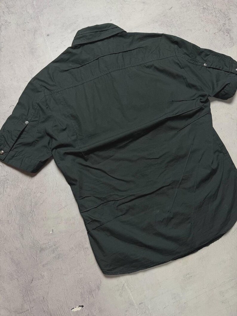 May include: Dark green short-sleeved shirt with a button-down collar. The shirt has two chest pockets and buttoned sleeve tabs. The back of the shirt is visible, showing seams and a curved hem. The fabric appears to be a lightweight material.