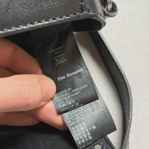 Puede incluir: Bolso de mano de cuero negro con herrajes plateados. La etiqueta dice "The Kooples" e incluye detalles como "COPYRIGHT KS14 AFS804 BLA01 C053934" e instrucciones de cuidado. El bolso tiene tachuelas.