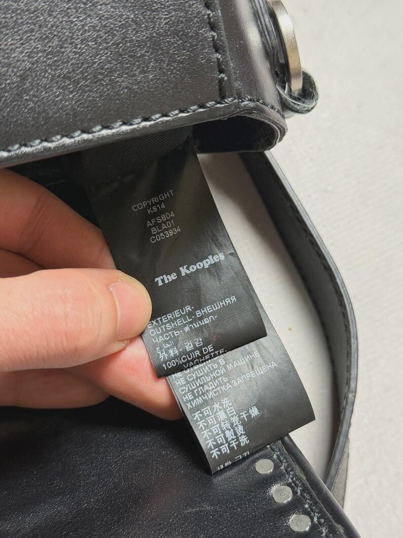 Puede incluir: Bolso de mano de cuero negro con herrajes plateados. La etiqueta dice "The Kooples" e incluye detalles como "COPYRIGHT KS14 AFS804 BLA01 C053934" e instrucciones de cuidado. El bolso tiene tachuelas.