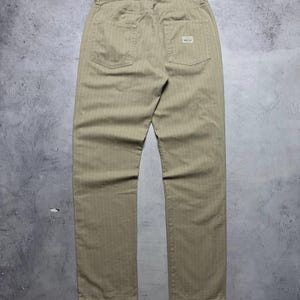Puede incluir: Pantalones color beige con finas rayas verticales. Los pantalones tienen dos bolsillos traseros y una pequeña etiqueta rectangular en el bolsillo trasero derecho. La tela parece resistente y la prenda es nueva.