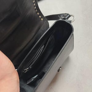 Puede incluir: Bolso bandolera de cuero negro con tachuelas plateadas en la solapa. El bolso tiene forma rectangular, un bolsillo interior con cremallera y un cierre plateado. La correa ajustable es negra con una raya blanca.