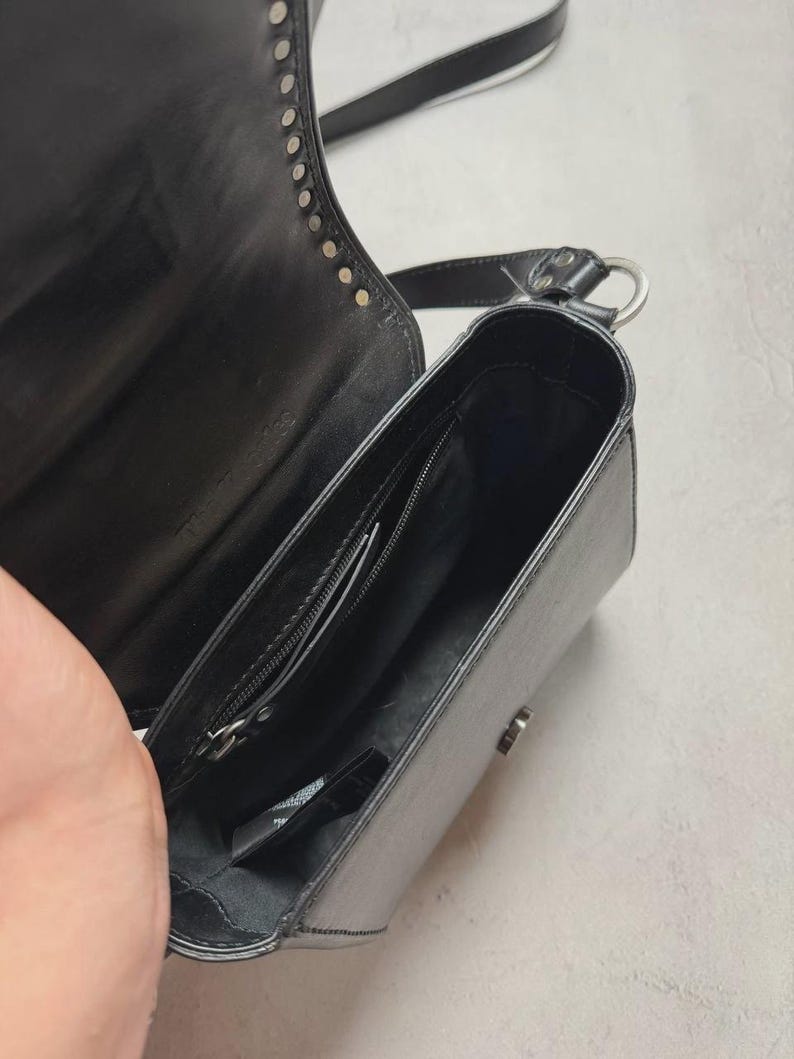 Puede incluir: Bolso bandolera de cuero negro con tachuelas plateadas en la solapa. El bolso tiene forma rectangular, un bolsillo interior con cremallera y un cierre plateado. La correa ajustable es negra con una raya blanca.