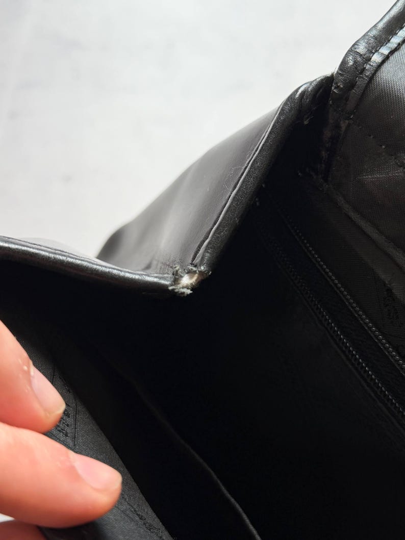 May include: Close-up of a black leather bag with a visible tear at the seam. The bag's interior is also visible, revealing a black fabric lining and a zipper. The image highlights the bag's texture and construction details.