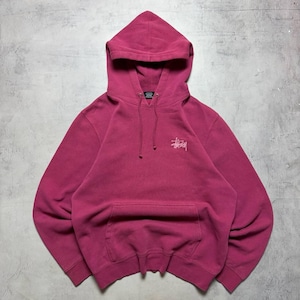 May include: A magenta long-sleeve hooded sweatshirt featuring a front kangaroo pocket, a drawstring hood, and a small white embroidered Stüssy logo on the left chest. The garment has ribbed cuffs and a ribbed hem, providing a structured fit.