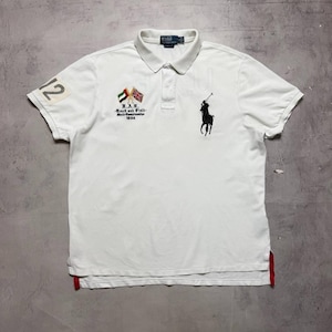 May include: A white short-sleeved polo shirt featuring a black embroidered polo player logo on the right chest and two crossed flags above the text U.A.E. Track and Field World Championships 1934. The right sleeve has a patch with the number 12.