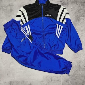Könnte beinhalten: Ein Vintage Adidas Trainingsanzug in Blau, Schwarz und Weiß. Die Jacke hat einen schwarzen oberen Bereich mit weißen Streifen auf den Ärmeln und dem Adidas-Logo. Die passende Hose ist blau mit dem Adidas-Logo. Der Trainingsanzug ist aus glänzendem, nylonartigem Material.