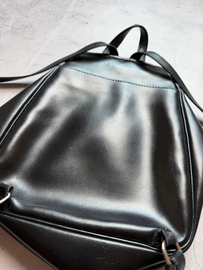 May include: A black, glossy backpack with adjustable straps and a top handle. The bag has a sleek, minimalist design, with a smooth, reflective surface. The backpack appears to be made of a durable material, suitable for everyday use.