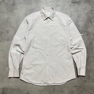 Pode incluir: Uma camisa rosa claro com botões e mangas compridas. A camisa tem uma gola clássica e punhos abotoados. O tecido tem um padrão de riscas verticais subtil. A camisa está estendida numa superfície cinzenta.