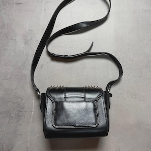 Puede incluir: Bolso bandolera negro con una correa larga y ajustable. El bolso tiene forma rectangular con cierre de solapa y tachuelas puntiagudas plateadas en el borde superior. El bolso est&aacute; hecho de un material liso y brillante.