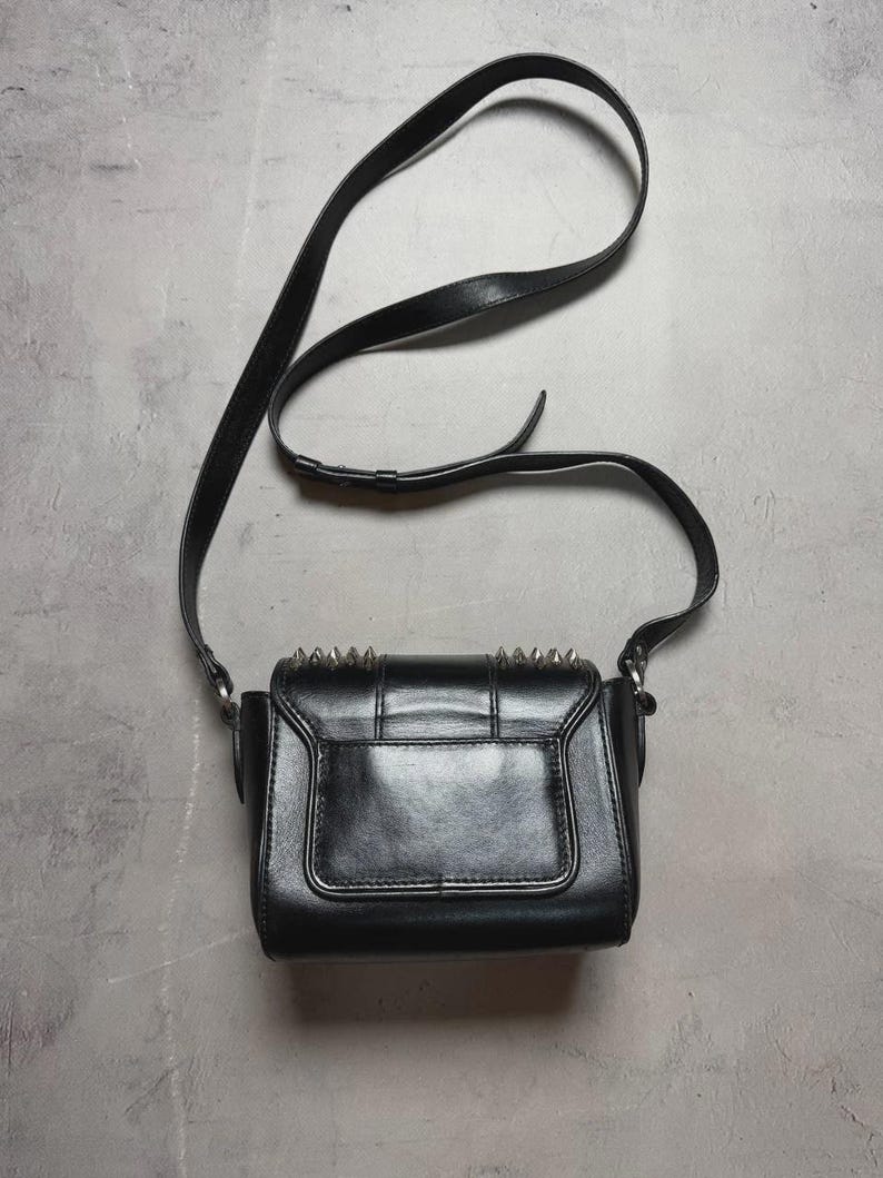 Puede incluir: Bolso bandolera negro con una correa larga y ajustable. El bolso tiene forma rectangular con cierre de solapa y tachuelas puntiagudas plateadas en el borde superior. El bolso est&aacute; hecho de un material liso y brillante.