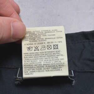 May include: A close-up of a clothing tag with text in multiple languages. The tag is white with black text and is attached to a dark fabric. The tag includes washing instructions and fabric composition.