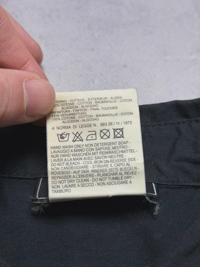 May include: A close-up of a clothing tag with text in multiple languages. The tag is white with black text and is attached to a dark fabric. The tag includes washing instructions and fabric composition.
