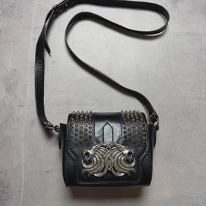 Puede incluir: Bolso bandolera de cuero negro con tachuelas de metal plateado y un emblema decorativo de cabeza de le&oacute;n. El bolso tiene una correa larga y ajustable y una forma rectangular.