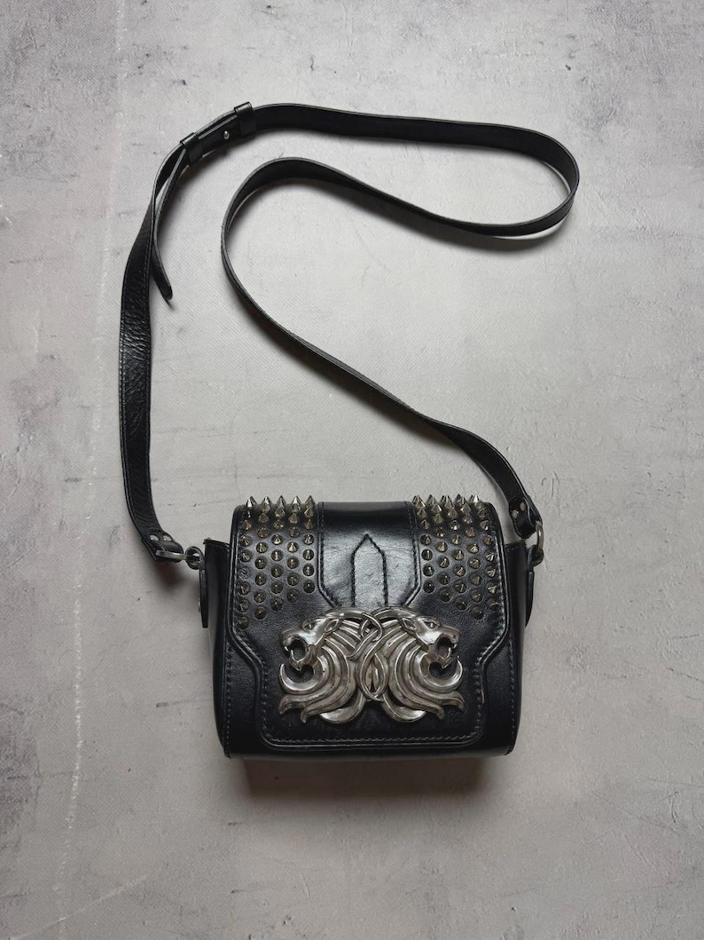 Puede incluir: Bolso bandolera de cuero negro con tachuelas de metal plateado y un emblema decorativo de cabeza de le&oacute;n. El bolso tiene una correa larga y ajustable y una forma rectangular.