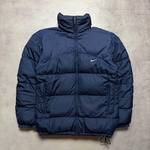 May include: A navy blue puffer jacket featuring a high collar, a full-length front zipper, and horizontal quilted panels. A small white Nike swoosh logo is embroidered on the upper left chest. The jacket includes two side pockets and a drawstring hem.