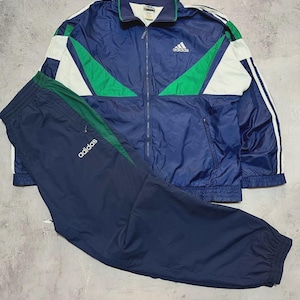 May include: A vintage Adidas tracksuit in navy blue, green, and white. The jacket has a zip-up front, a green collar, and white stripes on the sleeves. The matching pants have a green stripe down the side. The Adidas logo is visible on both pieces.