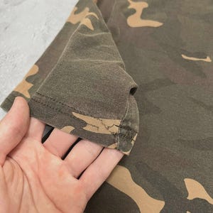 Puede incluir: Primer plano de una camiseta con estampado de camuflaje. La camiseta presenta una mezcla de colores verde oliva, marr&oacute;n y beige. La tela parece ser un material suave, posiblemente de algod&oacute;n. La manga es visible, con el borde de la manga enfocado.
